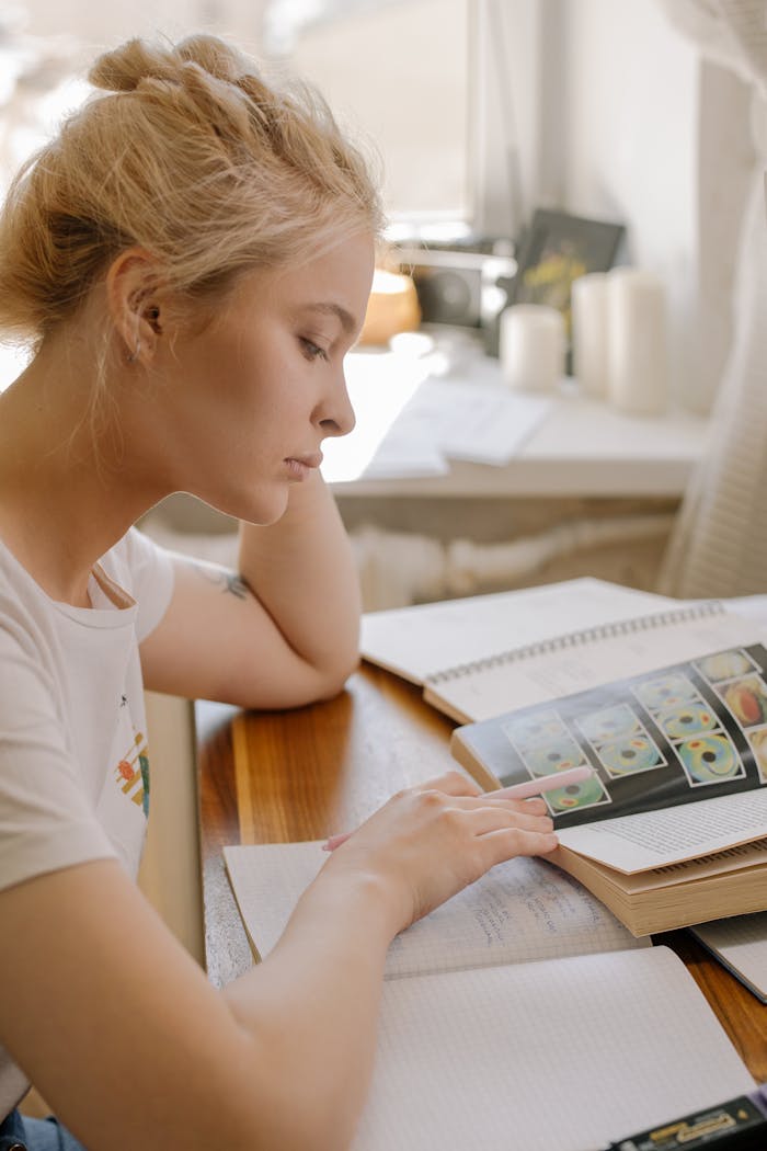 Focused young woman studying at home by the window with books open, illustrating a learning environment.